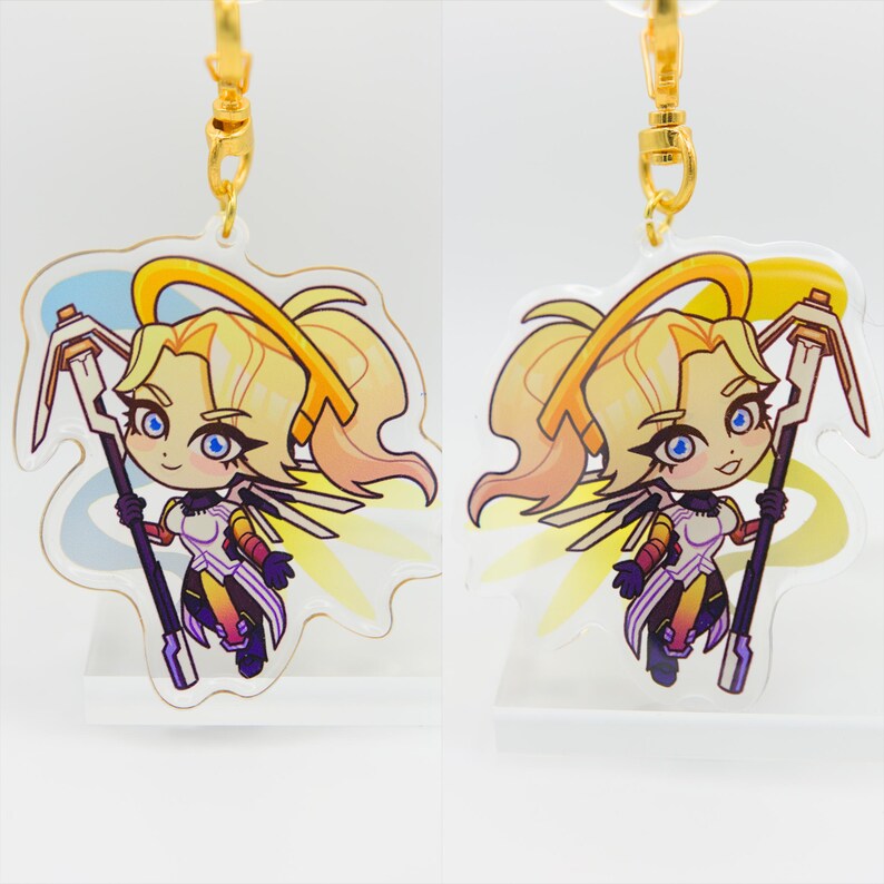 May include: A pair of gold keychains featuring a cartoon character with blonde hair, a white and yellow outfit, and a halo. The character is holding a staff with a blue and white design. The keychains are made of clear acrylic.