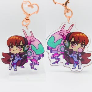 May include: A pink and green anime character keychain with a pink and green mech. The character has long brown hair and is wearing a purple and blue outfit. The keychain is attached to a gold metal chain.