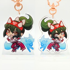 May include: A double-sided acrylic keychain featuring a cartoon character with green hair, red and white clothing, and a fox tail. The character is holding a peace sign with one hand and has a fox spirit behind them. The keychain is attached to a gold-colored chain.