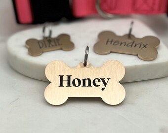 May include: Three gold bone-shaped dog tags with engraved names. The tags are Dixie, Hendrix, and Honey.
