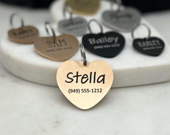 May include: A collection of gold, silver, and black heart-shaped dog tags with engraved names and phone numbers. The tags are arranged on a white marble surface. The tags read "Lily", "Sam", "Bailey", "Harley", and "Stella".