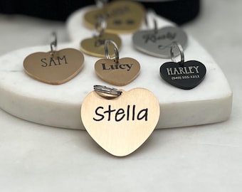 May include: Five gold and silver heart-shaped dog tags with engraved names and phone numbers. The tags are arranged on a white marble surface. The names on the tags are Sam, Lucy, Harley, Stella, and Rusty.