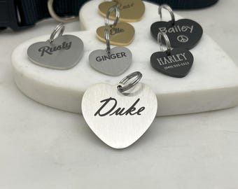 May include: A collection of heart-shaped dog tags with various names engraved on them, including Rusty, Elle, Ginger, Bailey, Harley, and Duke. The tags are made of silver and black metal and are displayed on a white surface.