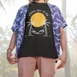 May include: A black t-shirt with a white graphic of a palm tree with a hammock and a yellow sun. The text on the shirt reads "#NOMAD, DON'T BE MAD."