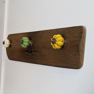 Peut inclure: Un porte-manteau mural en bois rustique avec trois boutons en céramique. Les boutons sont en forme de citrouille et peints dans des tons de vert, jaune et blanc.