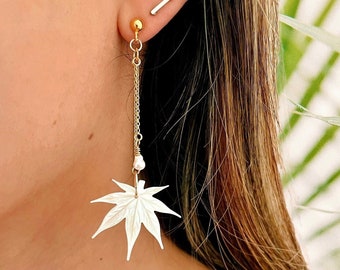 May include: A pair of gold earrings with a white maple leaf charm and a pearl bead. The earrings are dangling and have a delicate chain.