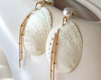 May include: Elegant dangle earrings featuring iridescent white seashell pendants, accented with gold-tone chains and a pearl stud. The earrings are designed with a delicate, feminine aesthetic, perfect for adding a touch of coastal charm to any outfit.