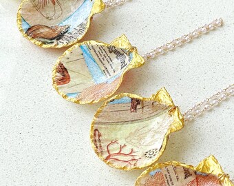 May include: Five gold-painted seashells with different vintage illustrations and text printed on them. Each seashell is attached to a delicate pearl necklace.