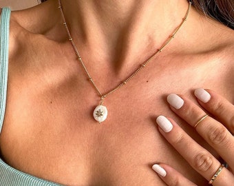 May include: A gold chain necklace featuring a small, white, irregular-shaped pearl pendant with a tiny gold star-shaped emblem centered on its surface. The necklace is worn with two gold rings on the fingers, one plain and one with small stones.
