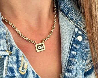 May include: A gold chain necklace with a square pendant featuring an eye design. The necklace is worn by a person wearing a denim jacket.