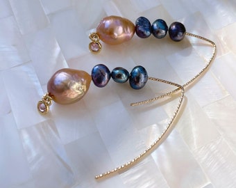 May include: A pair of gold earrings with a pink baroque pearl and three dark blue pearls. The earrings have a delicate chain design.