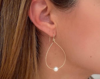 May include: A gold-toned teardrop-shaped earring featuring a single white pearl at the base, suspended from a thin wire hook. The earring wire has a textured, shimmering surface, and the pearl is round, smooth, and white in color.