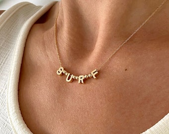 May include: Gold necklace with the word "SURF" spelled out in small letters. The necklace is on a model's neck.
