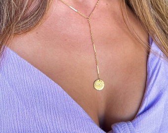 May include: A gold chain necklace with a small, round pendant featuring a detailed design. The necklace is layered, with a shorter chain at the top and a longer chain extending down to the pendant.