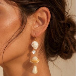 May include: Dangling earrings featuring natural seashells, a clear crystal, and a teardrop-shaped pendant. The earrings are suspended from gold-colored hooks. The shells have a cream and tan color palette.