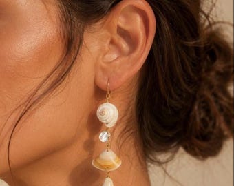 May include: Dangling earrings featuring natural seashells, a clear crystal, and a teardrop-shaped pendant. The earrings are suspended from gold-colored hooks. The shells have a cream and tan color palette.