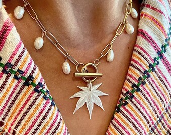 May include: A gold chain necklace with a white pearl pendant and a white mother-of-pearl leaf charm. The necklace is on a model wearing a striped shirt.