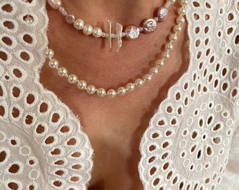 May include: Two layered pearl necklaces with a silver charm pendant. The top necklace features pink and white pearls, while the bottom necklace features white pearls.
