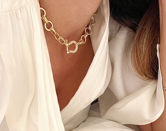 May include: A gold chain necklace with a large, round, pave-set clasp. The necklace is worn by a person with a white shirt.