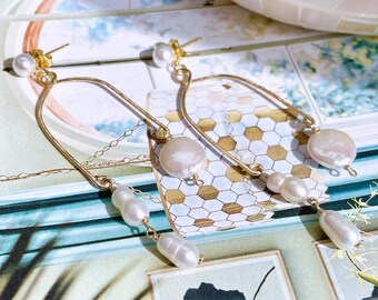 May include: Gold-toned, dangling earrings featuring lustrous, irregularly shaped white pearls. The earrings have a delicate, curved design with multiple pearls suspended from chains. The background includes a decorative tray and a patterned surface.