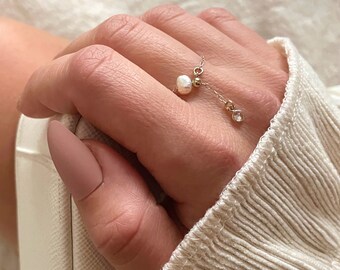 May include: A delicate gold chain ring with a pearl and a small clear crystal charm. The ring is worn on a finger with a light pink nail polish.