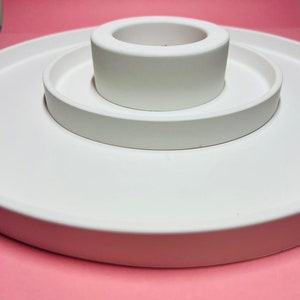 May include: A white ceramic tiered tray with three levels. The top level is a small round dish, the middle level is a larger round dish, and the bottom level is a wide, flat base.