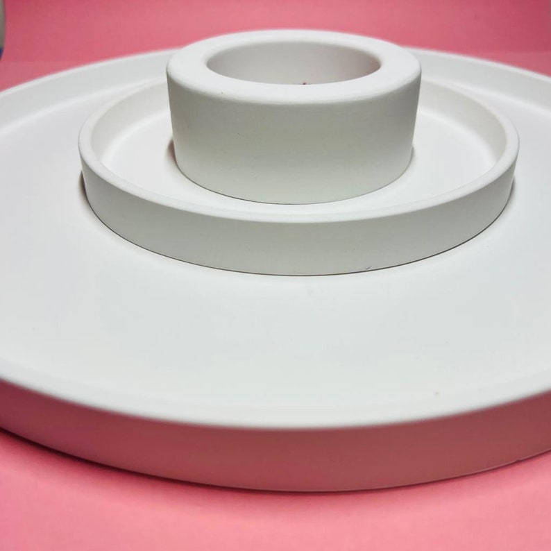 May include: A white ceramic tiered tray with three levels. The top level is a small round dish, the middle level is a larger round dish, and the bottom level is a wide, flat base.
