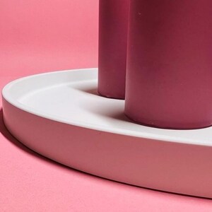 May include: Two tall pink candles on a white oval tray. The tray is on a pink background.