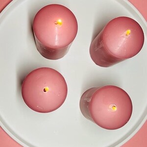May include: Four pink pillar candles with a single wick each, lit and burning on a white plate. The plate is on a pink surface.