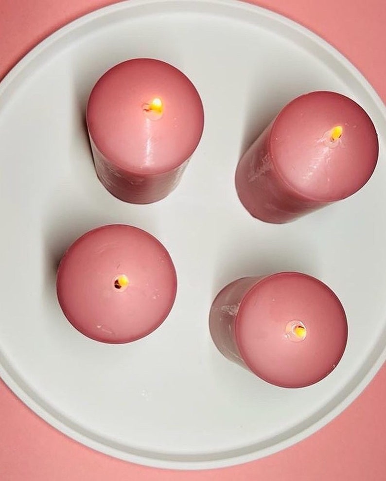 May include: Four pink pillar candles with a single wick each, lit and burning on a white plate. The plate is on a pink surface.