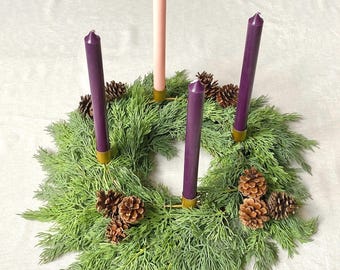 May include: An Advent wreath with four candles. Three purple candles and one pink candle are set in gold holders, surrounded by a green wreath with pinecones. The wreath is on a white surface.