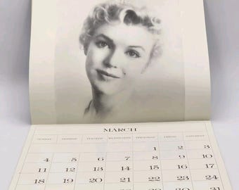 May include: A black and white calendar with the month of March displayed. The calendar features a photo of a woman with blonde hair. The woman is looking directly at the camera.