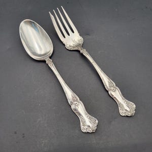 Puede incluir: Una cuchara y un tenedor de cuatro dientes en metal plateado con un patrón en relieve de uvas y hojas en los mangos. Ambos utensilios tienen un acabado pulido y un diseño decorativo a juego que se extiende desde la base del mango.