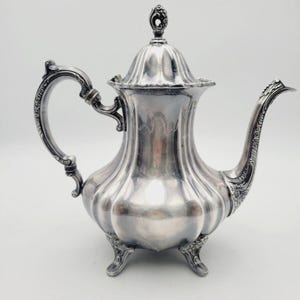 Könnte beinhalten: Eine silberne Teekanne mit geriffeltem Korpus, verziertem Henkel und drei Füßen. Die Teekanne hat einen Deckel mit einem dekorativen Knauf.