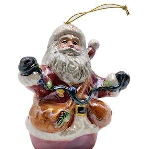May include: A ceramic Santa Claus Christmas ornament. Santa is wearing a red suit with white trim, holding a string of colorful lights. The ornament has a gold string for hanging and a detailed, hand-painted design.