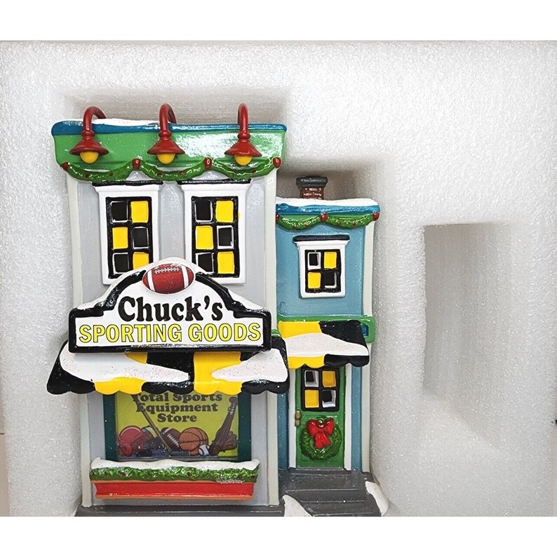 Pu&ograve; includere: Un modello in miniatura di un negozio di articoli sportivi chiamato "Chuck's Sporting Goods". L'edificio &egrave; dipinto in tonalit&agrave; di blu, verde e giallo con un tetto rosso. Il negozio ha un cartello con un pallone da calcio e una vetrina con attrezzature sportive.