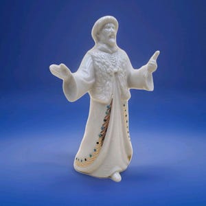 May include: A white porcelain figurine of a man in a long coat and hat, with arms outstretched. The coat is decorated with gold and teal accents. The figurine has a beard and is set against a blue background.
