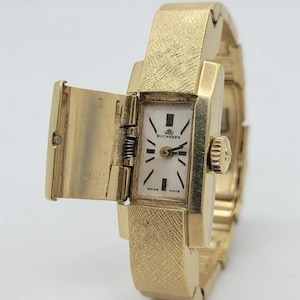 Puede incluir: Un reloj de pulsera Bucherer dorado con una esfera rectangular y una tapa con bisagras. La esfera del reloj es blanca con manecillas y marcadores negros, y se ven las palabras "Bucherer" y "Swiss Made". La correa del reloj tiene un acabado texturizado.