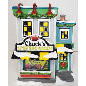 Pu&ograve; includere: Un edificio in miniatura con un tetto verde e un cartello che recita "Chuck's Sporting Goods". L'edificio ha un baldacchino bianco con strisce gialle e nere e una vetrina con attrezzature sportive. L'edificio &egrave; decorato con neve e una ghirlanda sulla porta.