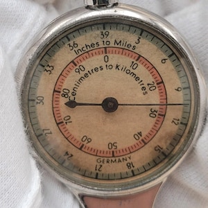 May include: A vintage silver map measurer with a circular face. The face displays measurements in inches to miles and centimetres to kilometres. The text "GERMANY" is visible at the bottom of the face.