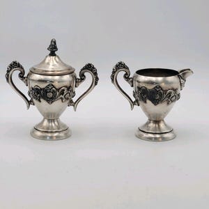 May include: A silver-colored sugar bowl with a lid and a creamer. Both pieces have ornate handles and decorative floral patterns. The sugar bowl has a small finial on top. The set is likely vintage or antique.