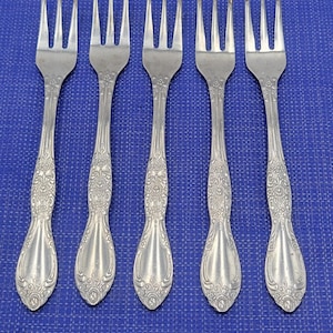 May include: Five silver-toned metal forks arranged in a row. Each fork features three tines and a handle decorated with an embossed floral pattern that extends from the neck down to the base, which terminates in a rounded, flared shape.