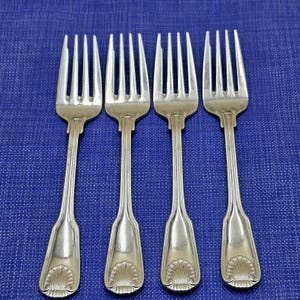 May include: Four silver forks with tines and shell-shaped designs on the handles are arranged on a blue surface. The forks are made of a shiny metal and have a classic, elegant design, suitable for formal dining.