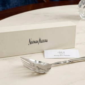 May include: A silver serving fork with a twisted handle, positioned in front of a rectangular off-white Neiman Marcus box and a small white card labeled Tuttle Silversmiths Sterling Silver Warranty. The fork has a polished, reflective finish.
