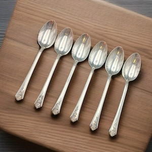 May include: Six silver spoons arranged on a wooden cutting board. The spoons have long, slender handles with decorative floral patterns near the bowl. The spoons are reflective, and the wooden board has a warm, brown tone.