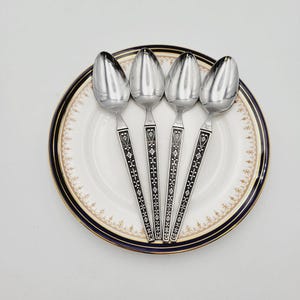 May include: Four silver spoons with ornate designs on the handles are arranged on a white plate with a blue and gold rim.