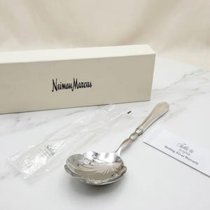May include: A silver-toned serving spoon with a shell-shaped bowl and a ribbed handle, accompanied by a white Neiman Marcus branded box, a clear plastic protective sleeve, and a small white card labeled Tuttle Sterling Silver Warranty.