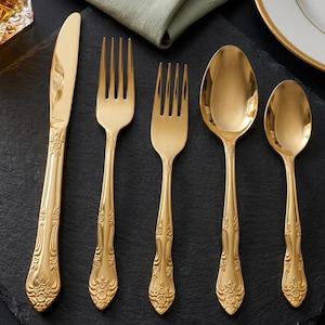 Puede incluir: Un juego de cinco cubiertos de tono dorado que consta de un cuchillo de mesa, un tenedor de mesa, un tenedor de ensalada, una cuchara de mesa y una cucharilla. Cada pieza presenta un acabado metálico pulido y un patrón floral en relieve.