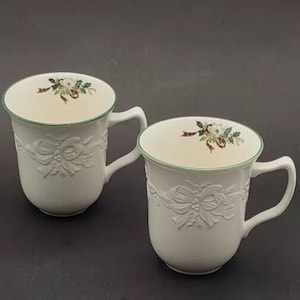 以下が含まれることがあります： 緑色の縁取りと花柄の白いセラミック製のマグカップ2つ。マグカップには、リボンとヒイラギのエンボス加工が施されています。