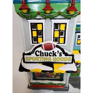 Pu&ograve; includere: Un edificio in miniatura in ceramica con un tetto verde e un cartello che recita "Chuck's Sporting Goods". L'edificio ha finestre gialle e un pallone da calcio sul cartello. L'edificio &egrave; decorato con una ghirlanda di foglie verdi e luci rosse.